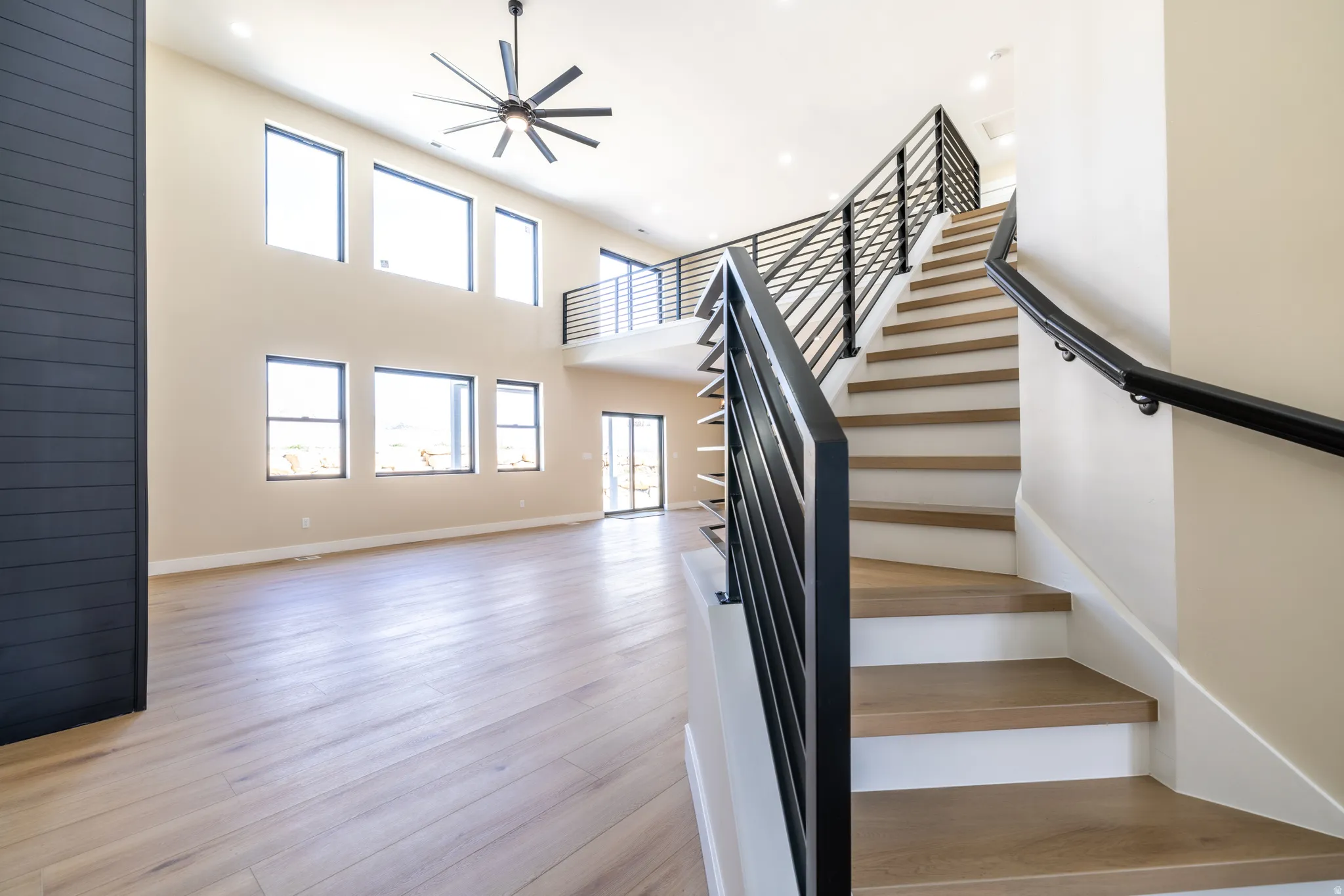 Two-story interior with wood-finish flooring, a contemporary ceiling fan, and a prominent staircase featuring black metal railings