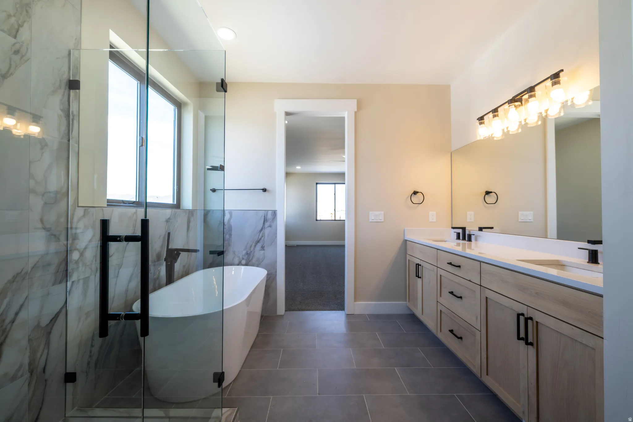 Freestanding soaking tub with a wall-mounted faucet, a glass-enclosed shower, a dual vanity with undermount sinks, and tile flooring