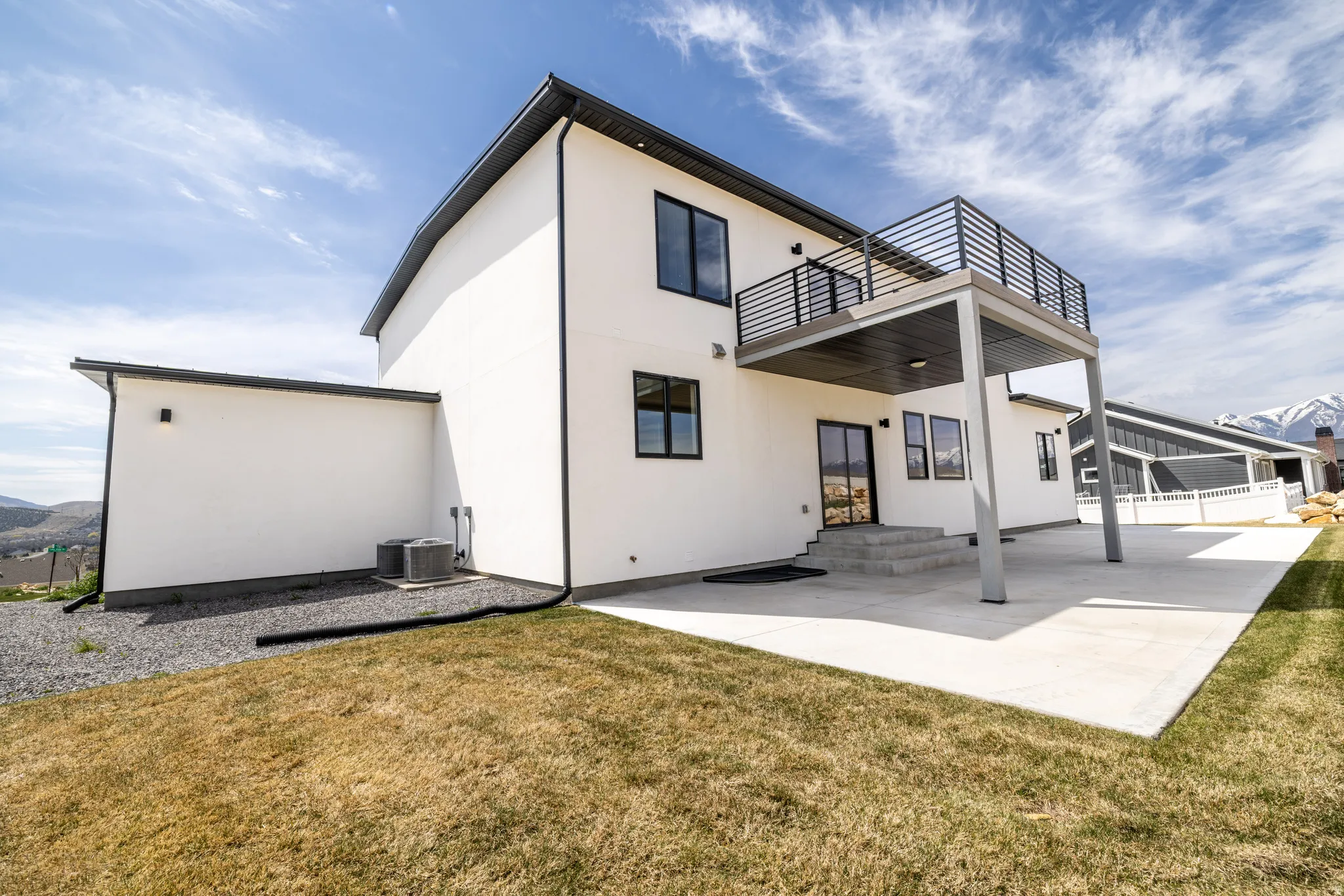 Contemporary exterior with white stucco finish, black trim accents, a second-story balcony with modern railing, a ground-level concrete patio, and a mountain backdrop