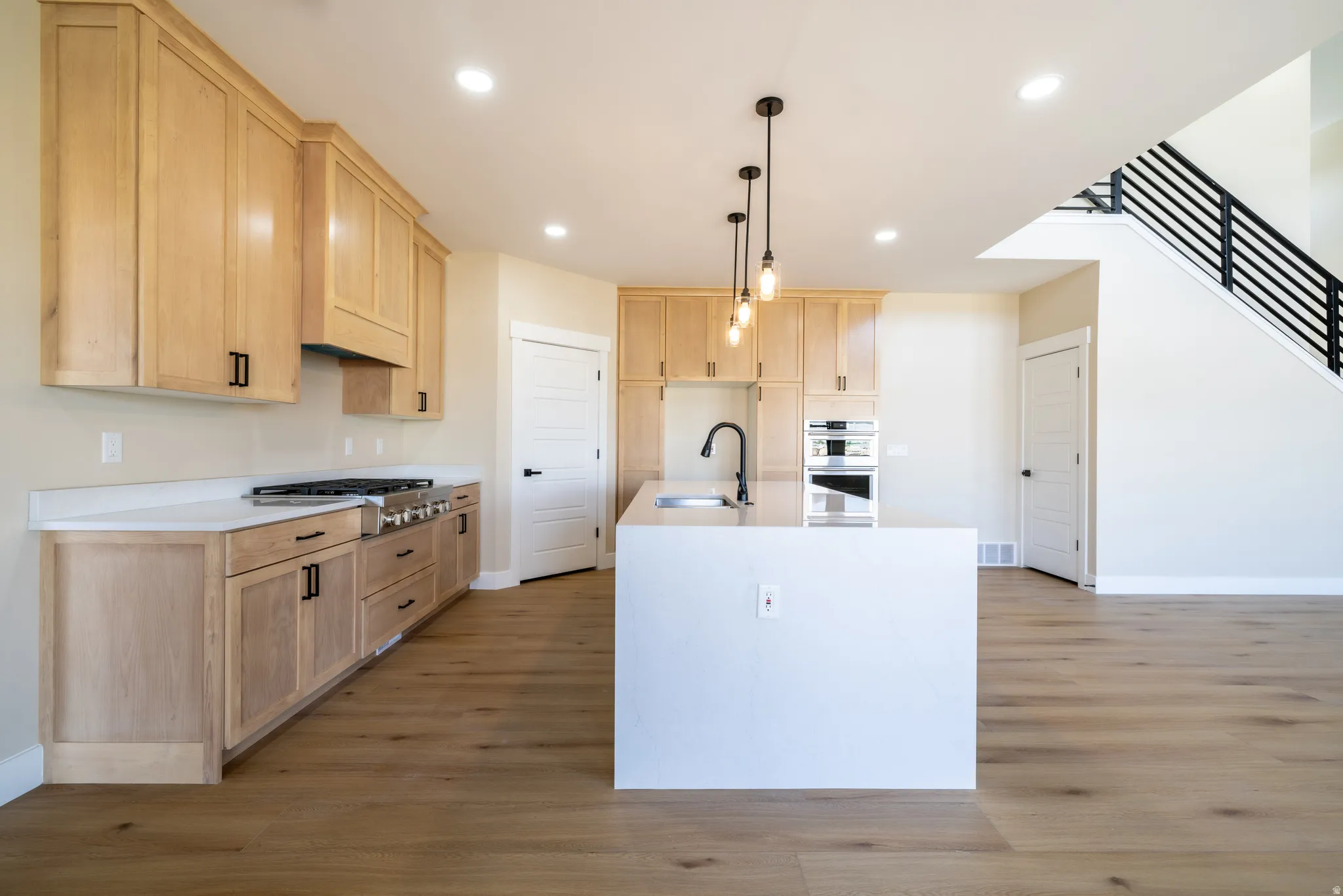 Spacious kitchen featuring light wood cabinetry, white countertops, built-in stainless steel appliances, a center island with a sink, and pendant lighting