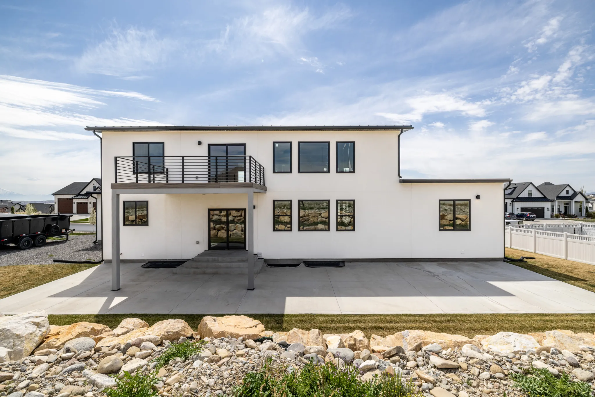 Contemporary exterior with white siding, black-framed windows, a second-story balcony with metal railing, and a ground-level concrete patio