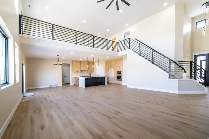 Open-concept living space featuring wood-finish flooring, a contemporary kitchen with light wood cabinetry and a dark island, and a prominent staircase with horizontal bar railings leading to an upper-level landing