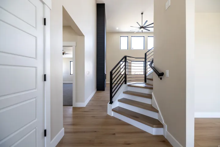 Light wood-finish flooring, two-story ceiling, black metal staircase railing, contemporary ceiling fan, and large windows