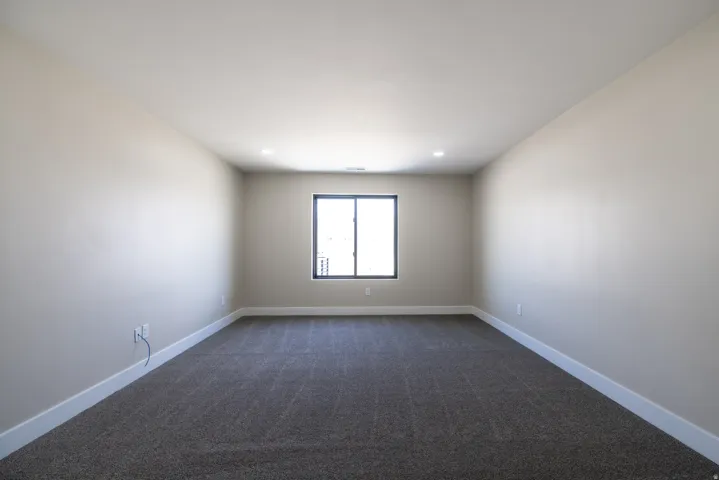 Expansive room with neutral tone walls, dark carpet, recessed lighting, and a single window with dark framing