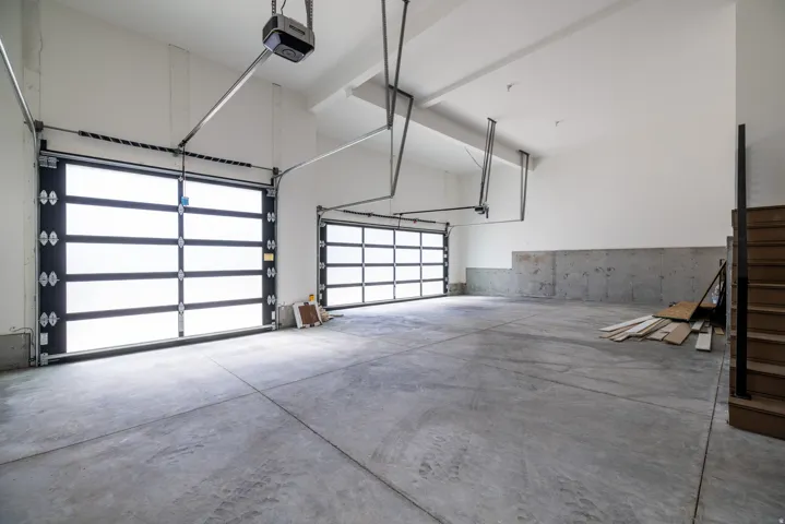 Spacious garage featuring two overhead doors with frosted glass panels, an exposed concrete floor, and a wooden staircase