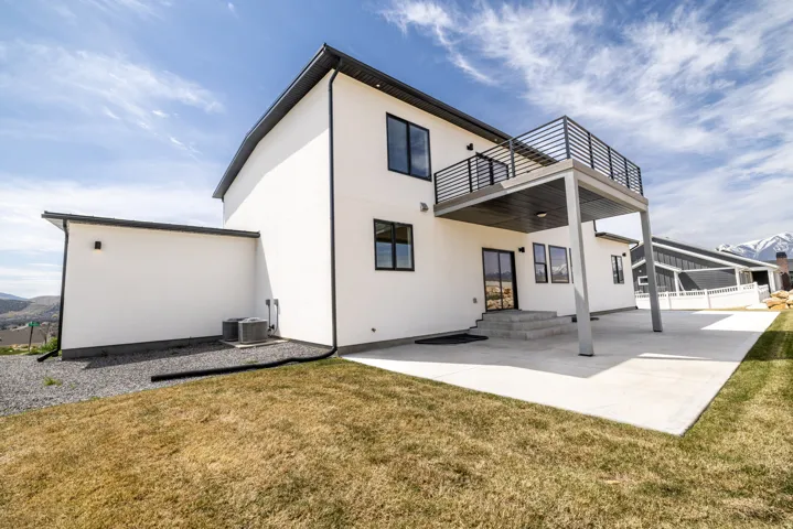 Contemporary exterior with white stucco finish, black trim accents, a second-story balcony with modern railing, a ground-level concrete patio, and a mountain backdrop