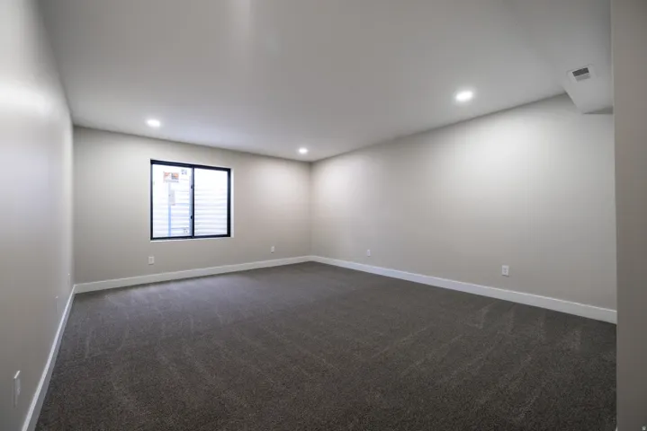 Recessed lighting, large window with blinds, light-toned walls, and dark patterned carpeting