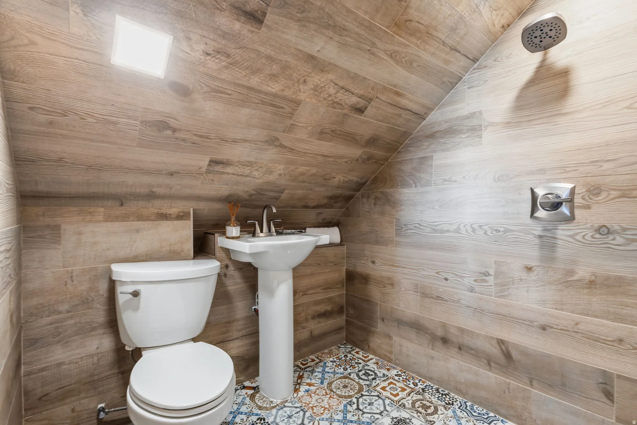 Wood-finish tile walls extending to an angled ceiling, pedestal sink with chrome faucet, rainfall showerhead, and patterned floor tiling