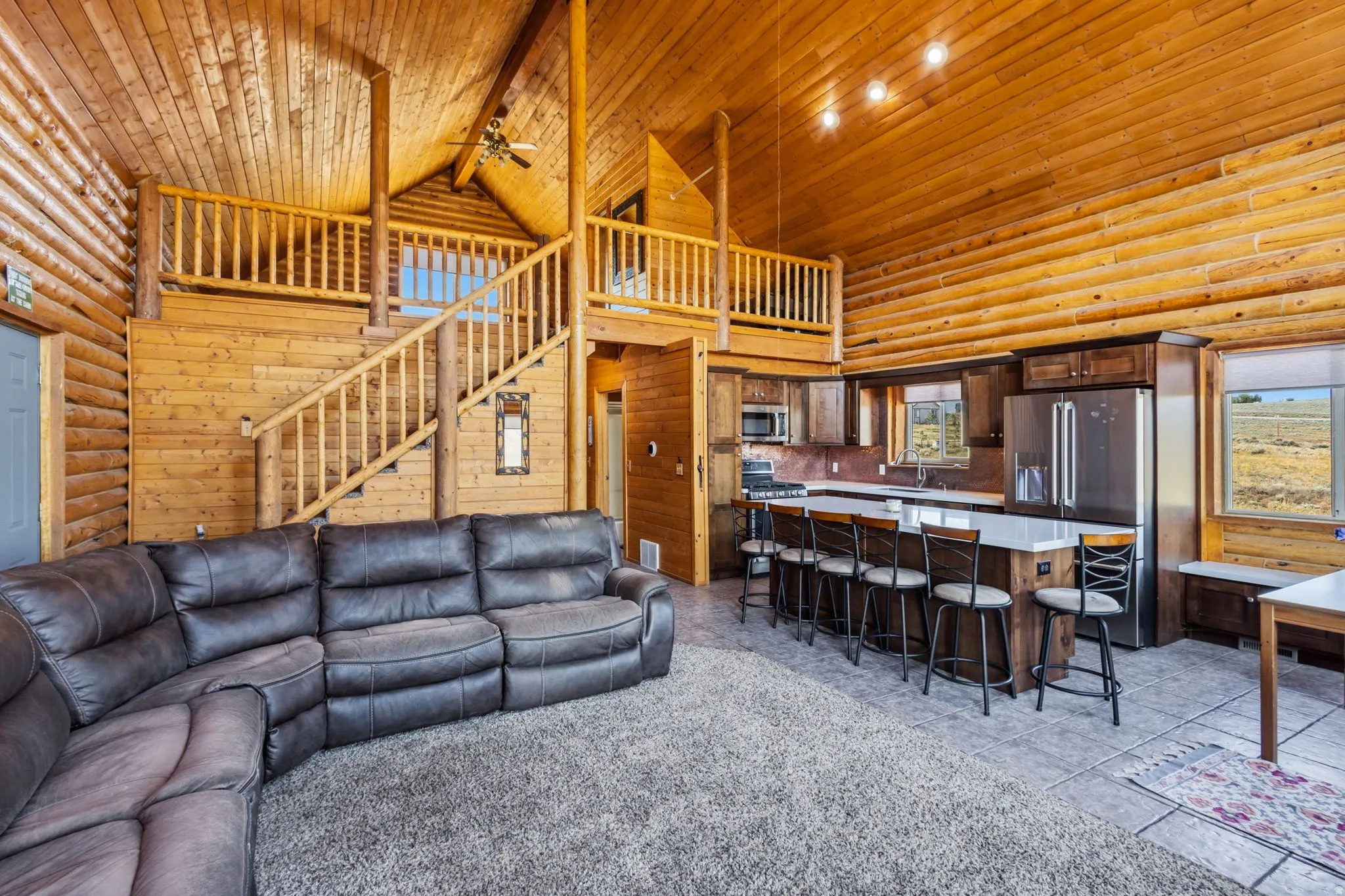 Exposed log walls with a vaulted wood-paneled ceiling, open loft area, and an integrated kitchen featuring stainless steel appliances and a counter-height island