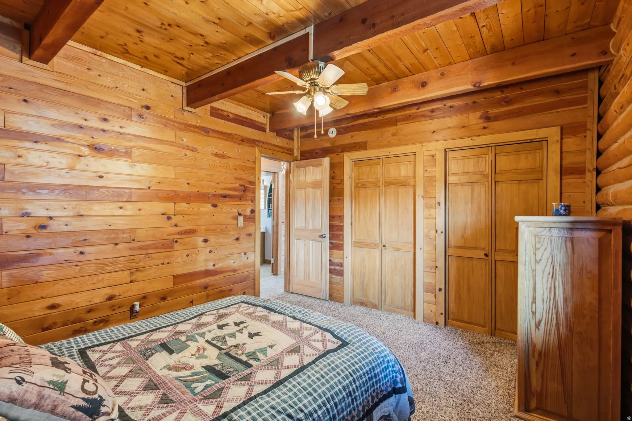 Wood-paneled walls and ceiling, exposed wood beams, ceiling fan with integrated lighting, two sets of bi-fold closet doors, and plush carpeting