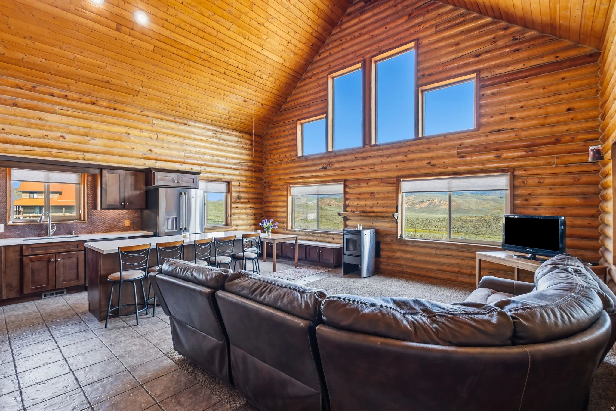 Vaulted wood-paneled ceiling with exposed beams, wood-plank walls, large picture windows, tile flooring, and a built-in kitchen island with seating