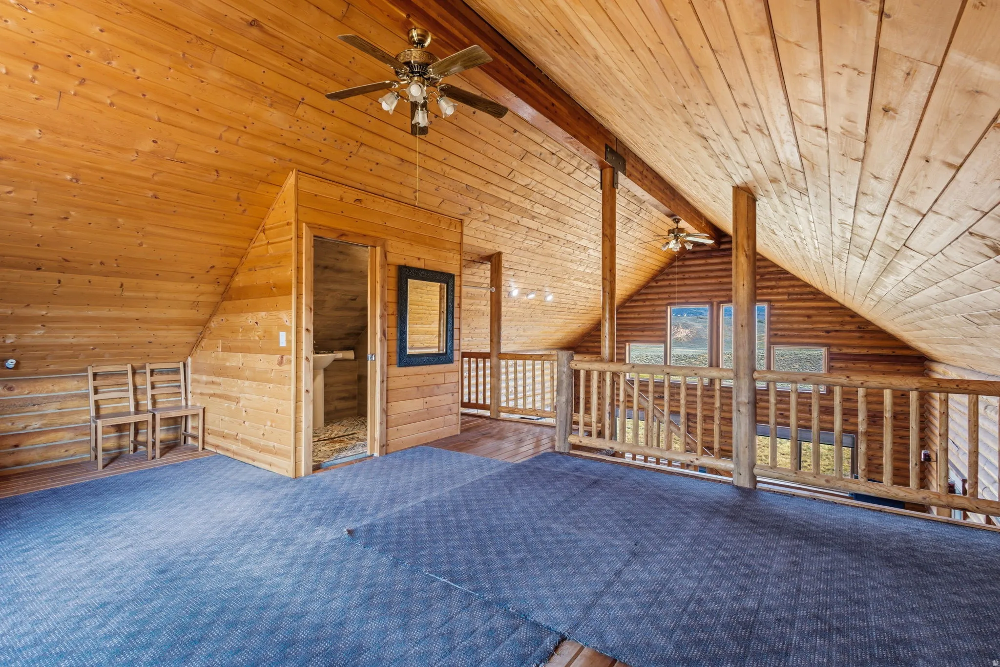 Vaulted wood-paneled ceiling, exposed wood beams, wood paneling throughout, ceiling fan with light fixture, and wood railing overlooking the lower level
