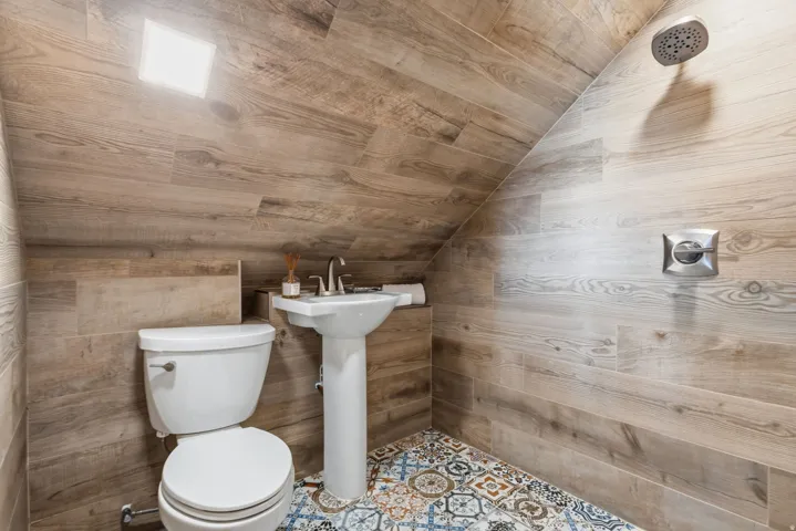 Wood-finish tile walls extending to an angled ceiling, pedestal sink with chrome faucet, rainfall showerhead, and patterned floor tiling