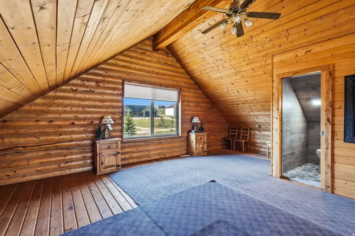 Wood-paneled sloped ceiling with exposed beams, wood-plank walls, window with roller shades, and a ceiling fan