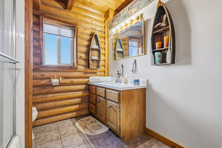 Rustic wood paneling, wood-finish vanity with dual sinks, white countertop, mirror with overhead lighting, and tiled flooring