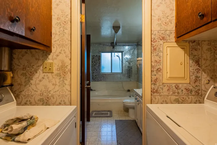 Galley-style laundry room featuring side-by-side washer and dryer hookups, wood-finish cabinetry with hardware accents, patterned wallcovering, and direct access to the adjoining bathroom with tile flooring and shower enclosure