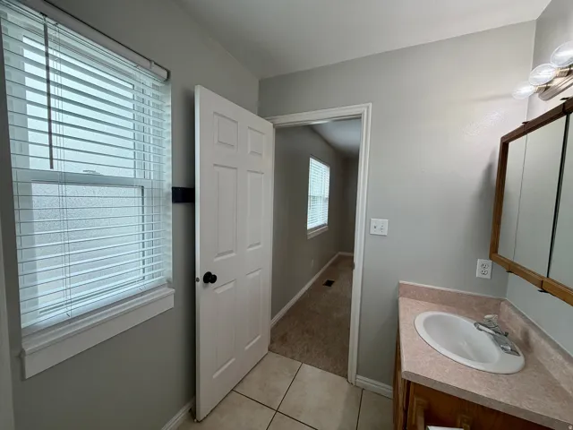 White single-pane window with horizontal blinds, tile flooring, wood-finish vanity with an integrated sink, and an overhead vanity light fixture