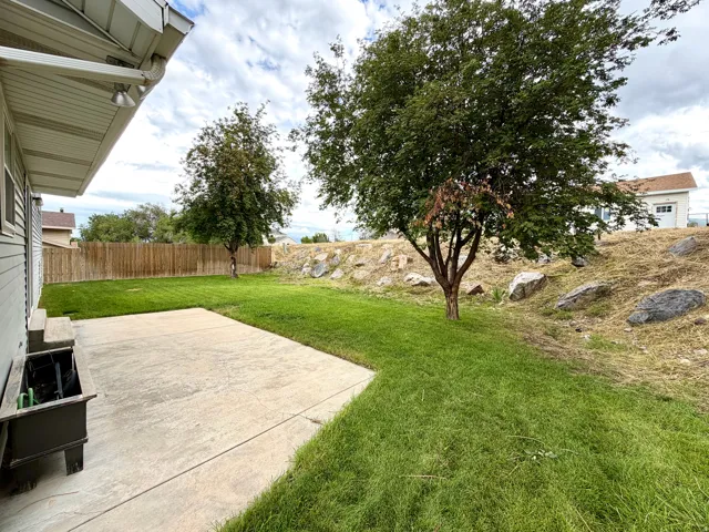 Concrete patio area, manicured lawn, full privacy fence, established trees, and natural rock landscaping