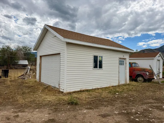 White vinyl-sided outbuilding with brown shingle roofing, featuring a roll-up garage door, a side entry door, and a single window