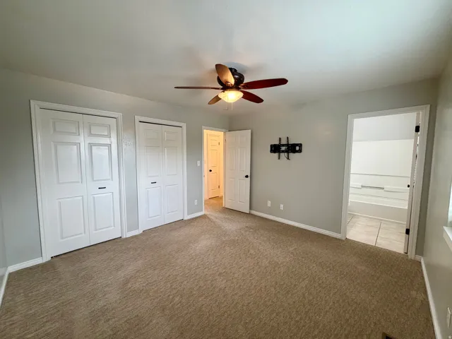 Expansive carpeted room featuring a ceiling fan, two double-door closets, and an open doorway to an adjacent room with tile flooring and a bathtub