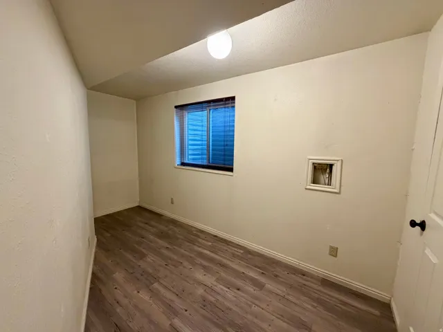 Wood-finish flooring with baseboard trim, a single window with blinds, and a built-in wall utility connection