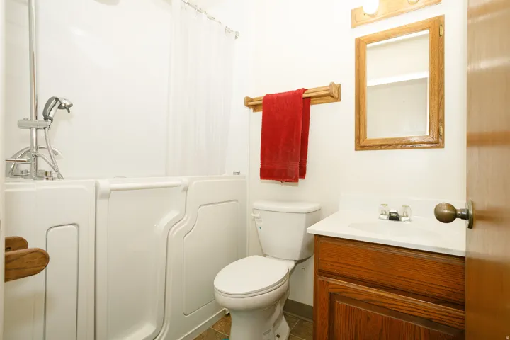 Walk-in tub with adjustable shower head, wood-finish vanity with integrated sink, wood-finish framed mirror, and tile flooring
