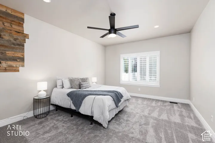 Accent wall featuring reclaimed wood paneling, recessed lighting, ceiling fan with matte black finish, window with white plantation shutters, and plush grey carpet