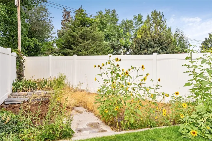 Private backyard retreat featuring mature landscaping, flowering perennials, stone pathway, lush green lawn, and white privacy fencing with mature trees visible beyond the property line
