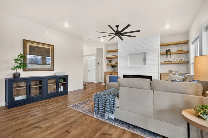 Wood-finish flooring throughout, complemented by a white shiplap fireplace with built-in shelving, a modern black ceiling fan, and recessed lighting
