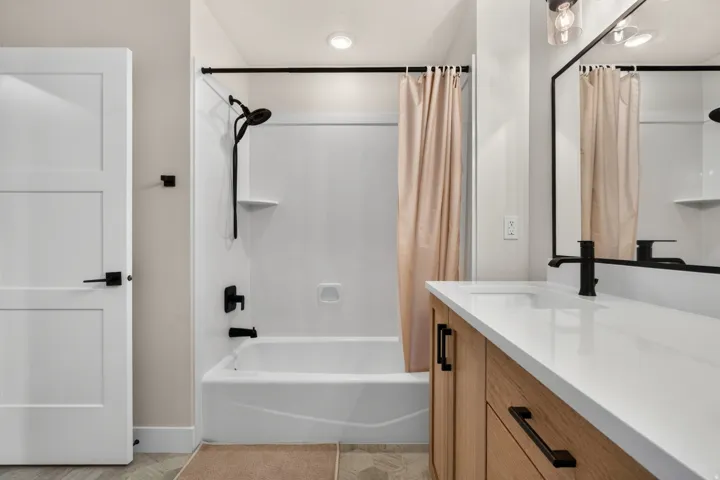 White three-panel door with black hardware, tub-shower combination with black fixtures, wood-finish vanity with white countertop, and rectangular black-framed mirror