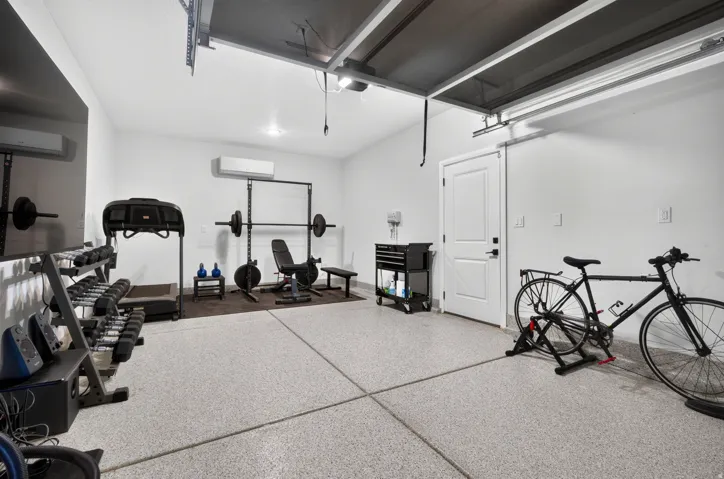 Dedicated fitness and recreation space featuring epoxy-coated modular flooring, overhead garage-style door system, recessed and pendant lighting, wall-mounted storage cabinetry, and ample room for cardio equipment, strength training apparatus, and recreat