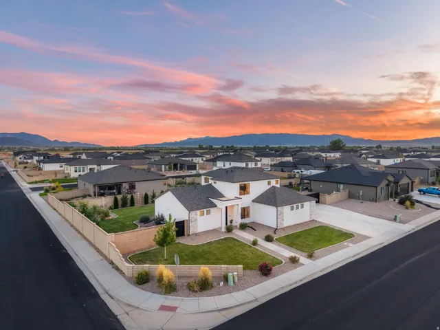 Modern two-story residence with white stucco and stone accents, featuring a multi-pitched dark roof, a corner lot with mature landscaping, a paved driveway, and an attached garage