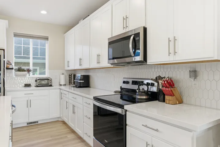 White cabinetry with brushed nickel hardware, light-toned wood-finish flooring, white countertops, stainless steel microwave, and geometric tile backsplash