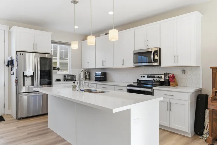 White shaker-style cabinetry, stainless steel appliances, a center island with sink, quartz countertops, and wood-finish flooring