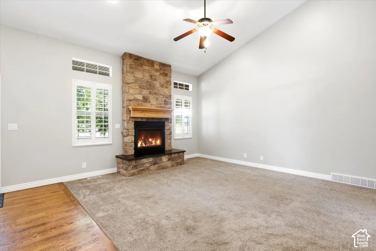 Vaulted ceiling featuring a ceiling fan, stone-clad fireplace with a wood mantle, windows with white plantation shutters, and wood-finish flooring