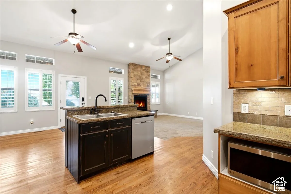 Hardwood flooring, kitchen island with sink, built-in stainless steel dishwasher, stone-clad fireplace, and vaulted ceilings