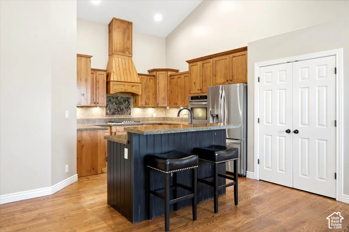 Kitchen featuring wood-finish flooring, natural wood cabinetry, a granite countertop island with a paneled base, stainless steel appliances, and a vaulted ceiling