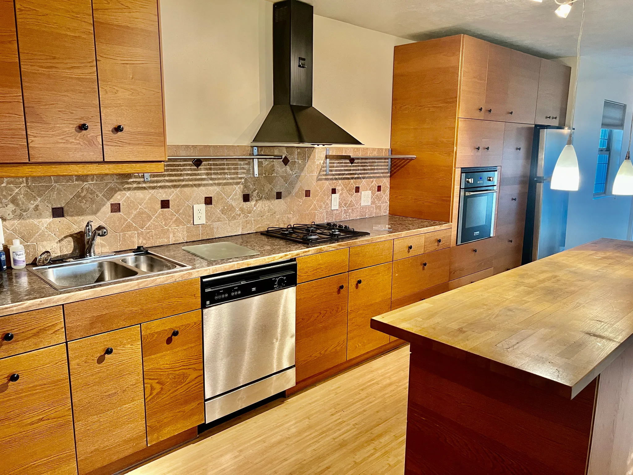 Wood-finish cabinetry with black hardware, stainless steel sink, built-in dishwasher, and a gas stovetop with overhead range hood