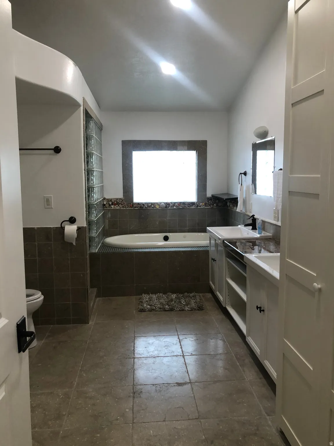 Tiled flooring throughout, soaking tub, dual vanity with undermount sinks, built-in cabinetry, and glass block shower surround