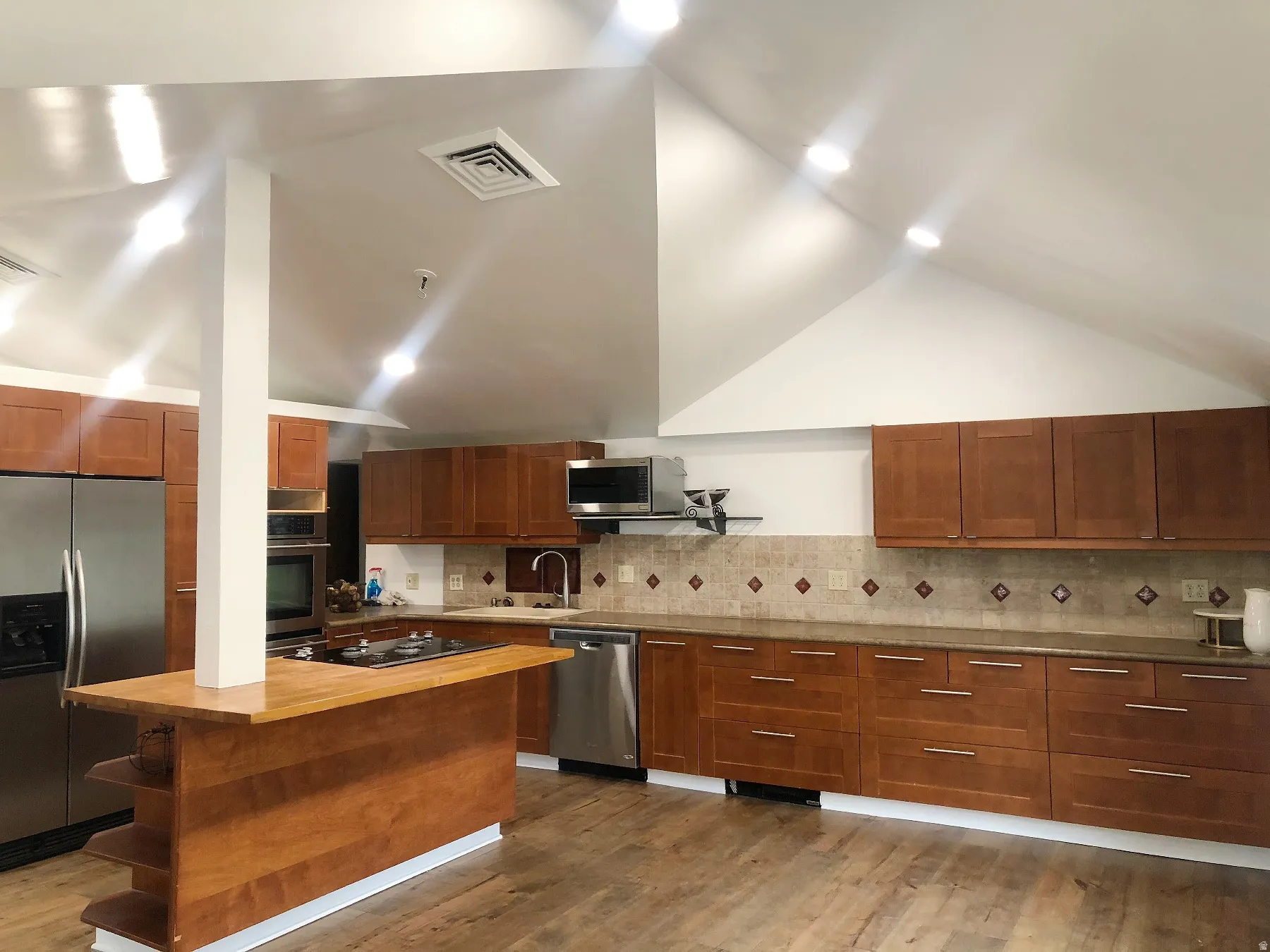 Vaulted ceiling with recessed lighting, wood-finish flooring, wood cabinetry, built-in stainless steel appliances, and a tiled backsplash