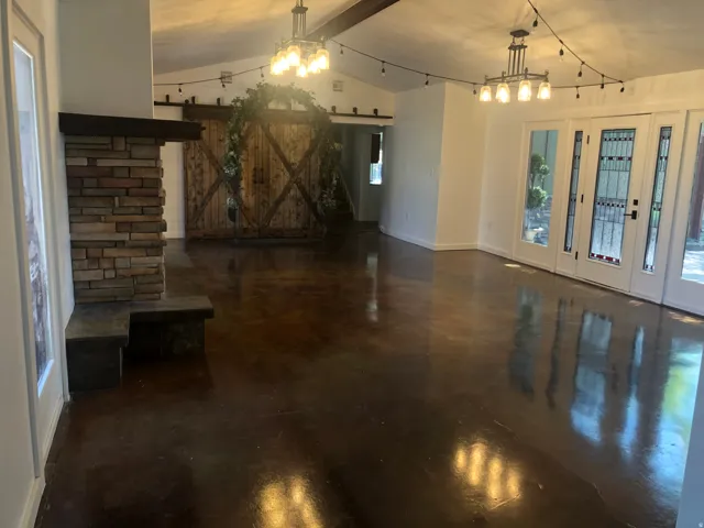 Exposed beam ceiling with vaulted design, stone-clad fireplace with hearth, polished concrete flooring, barn-style sliding doors, and double French doors with transom windows