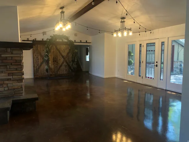 Vaulted ceiling with exposed wood beam, polished concrete flooring, stone-clad fireplace, and French doors with decorative glass inserts