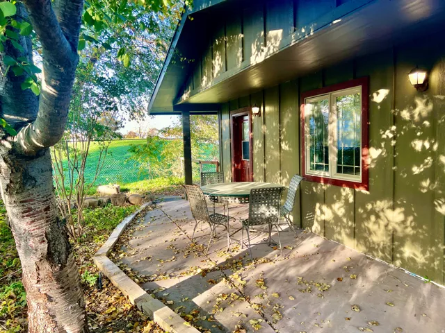 Exterior siding, covered patio, wall-mounted light fixtures, single window with red trim, and a prominent tree