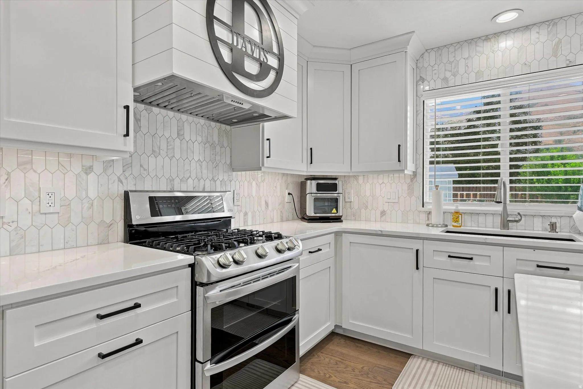 White shaker cabinetry, stone countertops, full-height backsplash with picket tile, stainless steel gas range, and wood-finish flooring