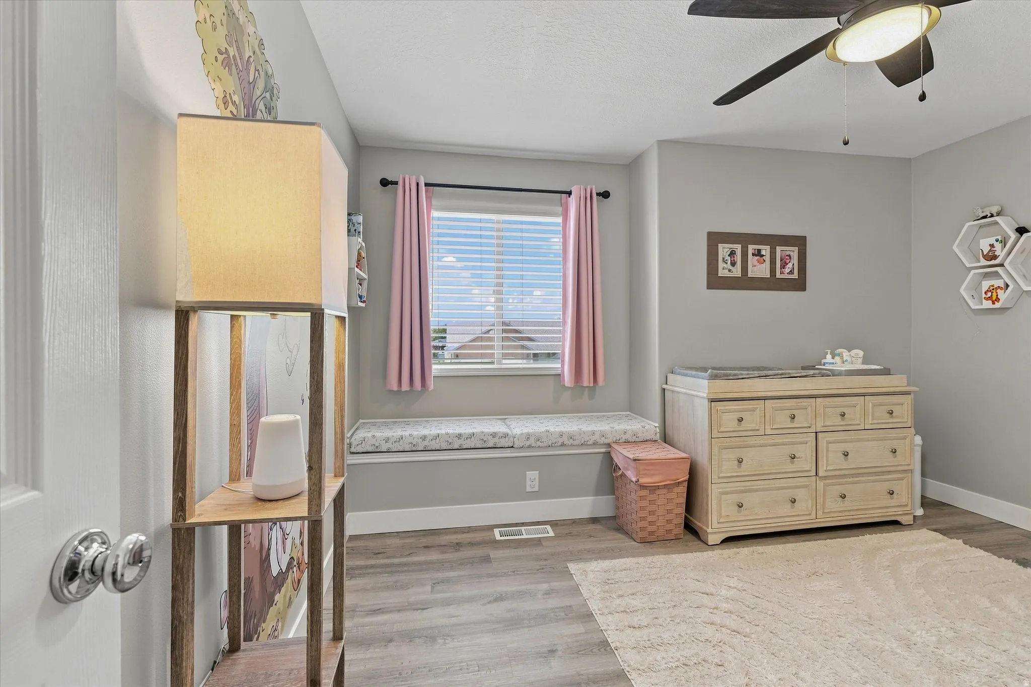 Window-mounted blinds with pink curtains, built-in window seat with cushion, overhead ceiling fan with lighting, wood-finish flooring, and light gray wall paint