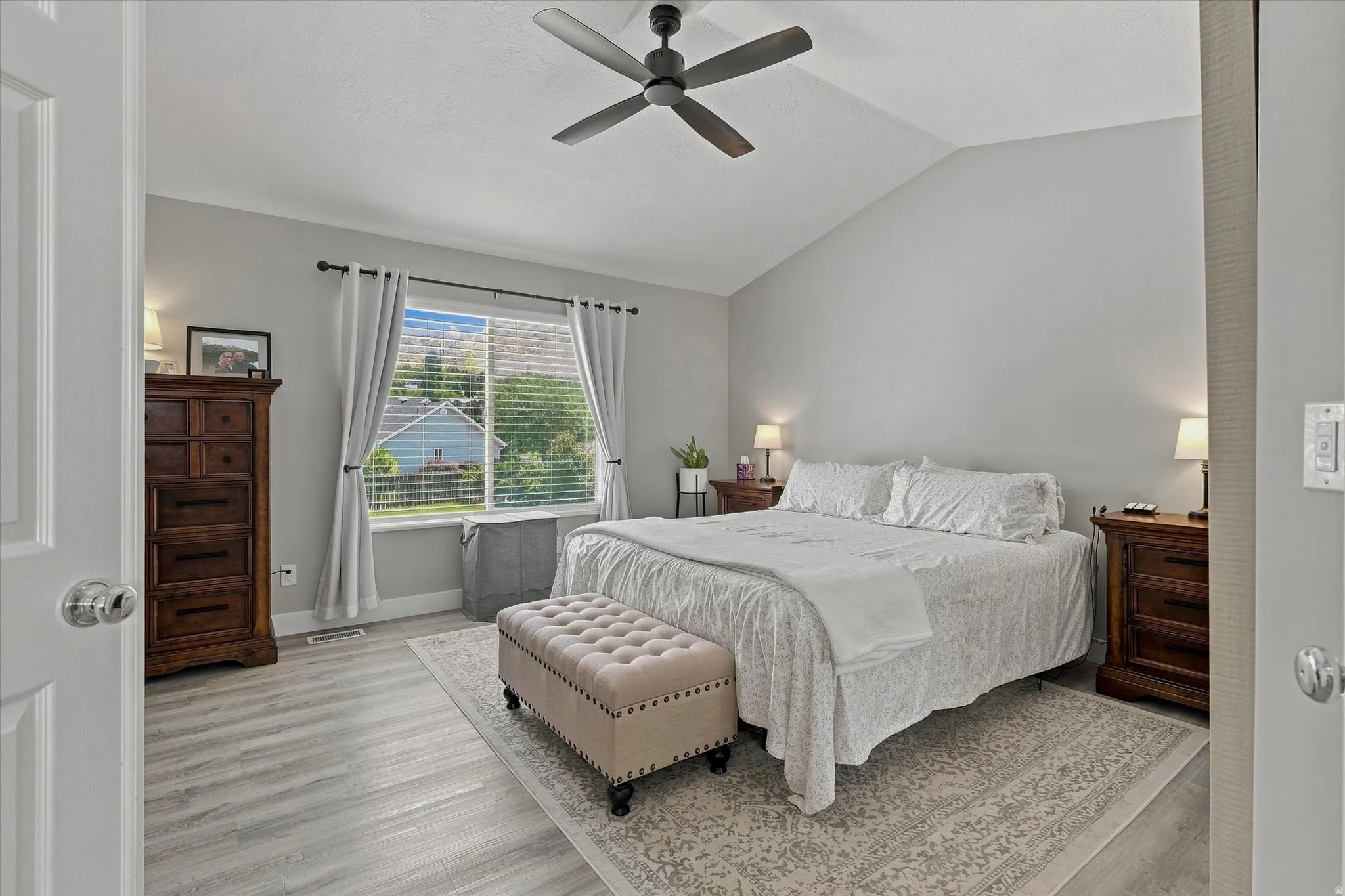 Vaulted ceiling, wood-finish flooring, ceiling fan, and a large window with blinds