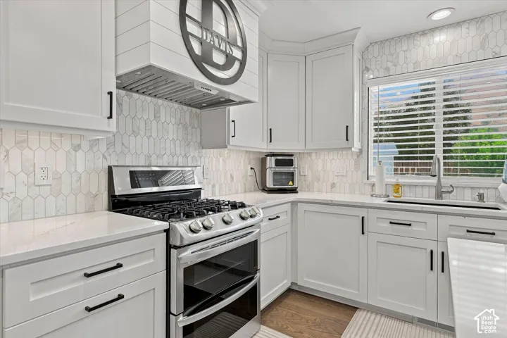 White shaker cabinetry, stone countertops, full-height backsplash with picket tile, stainless steel gas range, and wood-finish flooring