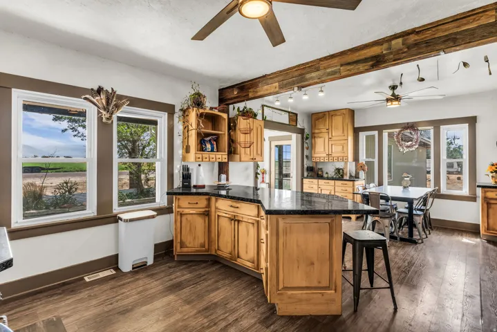 Wood-finish flooring, abundant natural light from multiple windows, dark countertops, wood cabinetry, and exposed wood beam accents