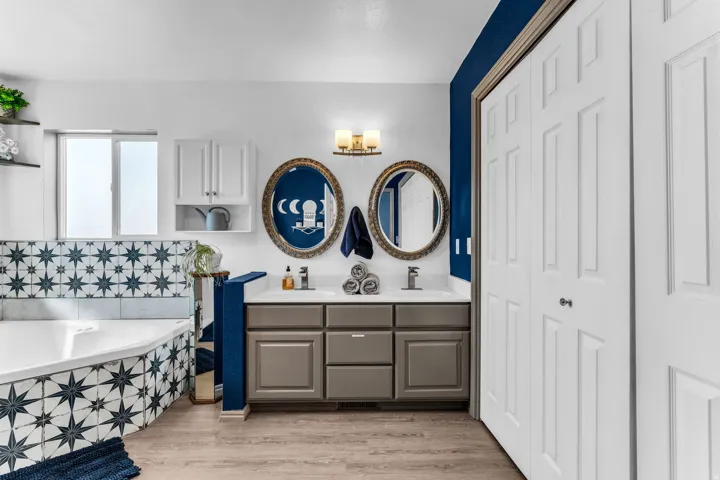 Dual vanity with individual oval mirrors, integrated corner soaking tub with patterned tile surround, wood-finish flooring, and white bifold closet doors
