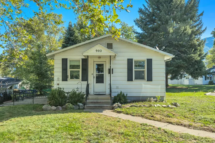 White exterior siding with black window shutters, concrete front walkway, metal porch railing, and a mature front yard tree